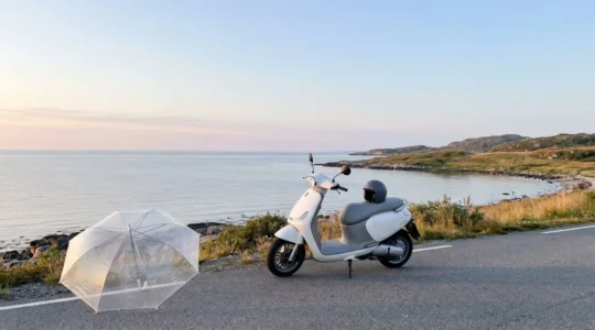 Scooter moderne garé sur route côtière au lever du soleil avec parapluie transparent au premier plan symbolisant la protection d'assurance